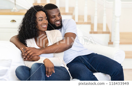 A man and woman are sitting on a white couch in a home. The man has his arm around the woman and they are both looking to the side. A man and woman are sitting on a white couch in a home. The man has his arm around the woman and they are both looking to the side. 118433949