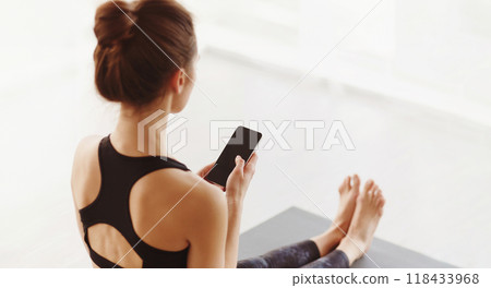 A woman is seated on a yoga mat indoors, engrossed in her cell phone. She appears focused as she browses, texting, or checking emails while in a cross-legged position. 118433968