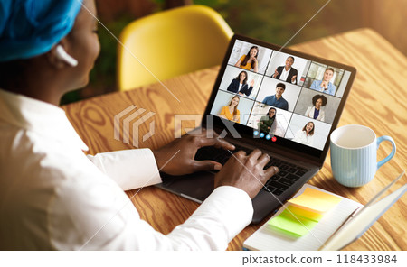 African American woman wearing a blue headband and white shirt sits at a wooden table, using a laptop to participate in a video conference meeting, cafe interior 118433984