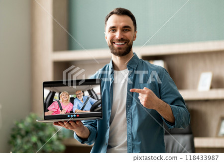 A man stands in a home office, smiling and pointing at his laptop screen, which shows a video call with an elderly couple sitting in a car. A man stands in a home office, smiling and pointing at his laptop screen, which shows a video call with an elderly couple sitting in a car. 118433987