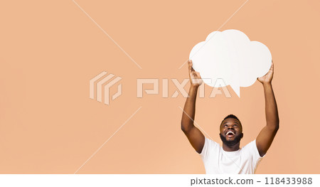 Happy Black man holding a blank speech bubble up above his head with his arms raised. He is looking up at the bubble and smiling broadly. The background is a simple peach color, copy space 118433988