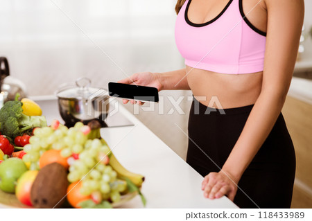 A woman in workout clothes holds a smartphone in a kitchen with fresh fruits and vegetables in the foreground. A woman in workout clothes holds a smartphone in a kitchen with fresh fruits and vegetables in the foreground. 118433989