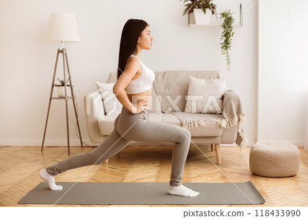 A woman in athletic wear does lunges on a yoga mat in a living room. A woman in athletic wear does lunges on a yoga mat in a living room. 118433990