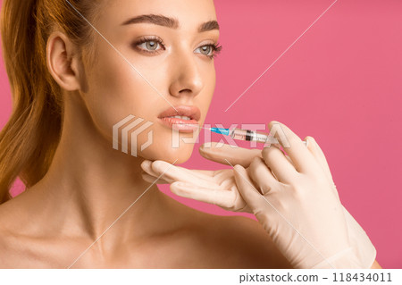 A young woman with long red hair is receiving lip injections from a medical professional wearing white gloves. The woman is looking to the side and the gloved hand holding the syringe 118434011