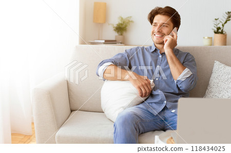 A man in a blue shirt sits on a white couch, smiling as he talks on his phone. A man in a blue shirt sits on a white couch, smiling as he talks on his phone. 118434025