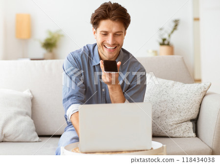 A man is sitting on a couch, smiling while looking at a smartphone in his hand. He appears to be using a voice assistant to complete a task while working on a laptop. A man is sitting on a couch, smiling while looking at a smartphone in his hand. He appears to be using a voice assistant to complete a task while working on a laptop. 118434038