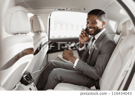 African American successful businessman, dressed in a suit, sits in the backseat of a car. He is talking on his phone, smiling, and giving a thumbs up. The car is clean and luxurious 118434043