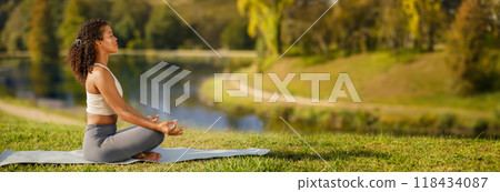 Side view of millennial woman in sports outfit meditating on yoga mat at lake in green park outside. Lady practicing breathing exercises in nature for inner peace and harmony outdoor, copy space 118434087