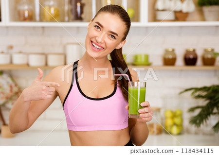 A woman in athletic wear smiles and holds a green smoothie in a glass with a straw. A woman in athletic wear smiles and holds a green smoothie in a glass with a straw. 118434115