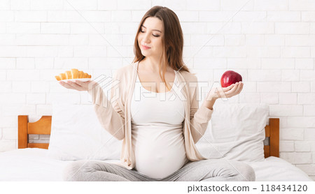 Healthy nutrition and pregnancy. Pregnant woman choosing between croissant and apple, sitting on bed at home 118434120