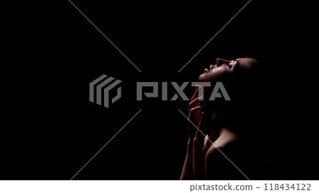A woman with her head tilted back, looking towards the top of the frame, is silhouetted against a dark background. She has her fingers gently on her neck, copy space 118434122