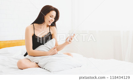 A woman is seated on a bed, holding a smartphone in her hands and looking at the screen. She appears focused on the device while sitting in a bedroom, copy space 118434129