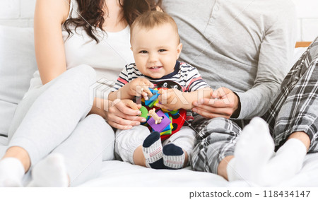 A happy baby sits on a bed with its parents, playing with a colorful toy. The baby is wearing a striped shirt and has a big smile on its face. Parents are looking at the baby with love and affection. 118434147