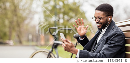 An African American businessman is on his phone, seated on a park bench with his bicycle near. The scene captures a moment of urban life in a green, peaceful environment. An African American businessman is on his phone, seated on a park bench with his bicycle near. The scene captures a moment of urban life in a green, peaceful environment. 118434148