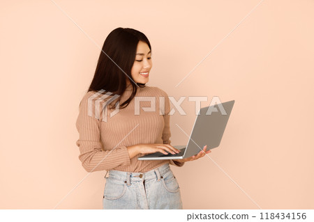 A young woman with long black hair smiles as she uses a laptop against a peach-colored background. 118434156