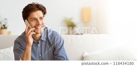 A man sits on a couch while talking on his phone, smiling and looking to his left. A man sits on a couch while talking on his phone, smiling and looking to his left. 118434180