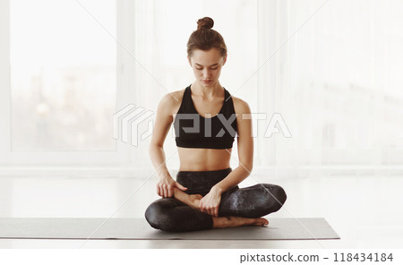 A young woman is seated on a yoga mat in a peaceful, well-lit room. She appears to be in deep concentration, maybe in meditation or preparing for a yoga session 118434184