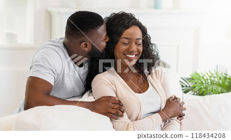 A man whispers to a woman on a white couch, she smiles and closes her eyes. 118434208