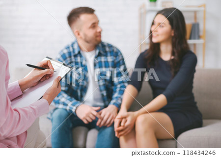 A therapist takes notes on a clipboard during a counseling session with a couple. The couple is sitting on a couch, holding hands, and listening intently to the therapist 118434246