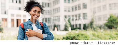 Portrait Of Successful Student. Happy African American Teenager Girl Posing Holding Books Smiling Looking At Camera Standing Near Modern University Building. Modern Education, Copy Space 118434256