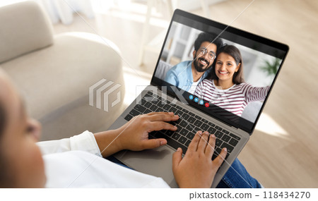 Black lady person sits on a couch at home, using a laptop computer to video chat with a couple. The person is typing on the laptop, while the couple smiles and waves at the camera. 118434270