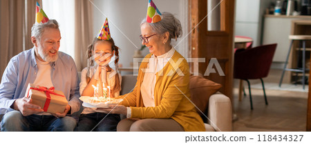 Three family members gather in a cozy living room to celebrate a birthday. The girl lights candles on a cake while the grandparents look on with joy, creating a heartwarming moment. 118434327
