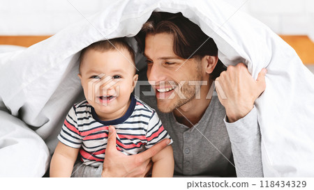 A father and his baby son are lying in bed, peeking out from under a white blanket. They are both smiling, and the father is holding his son. They appear to be having fun 118434329