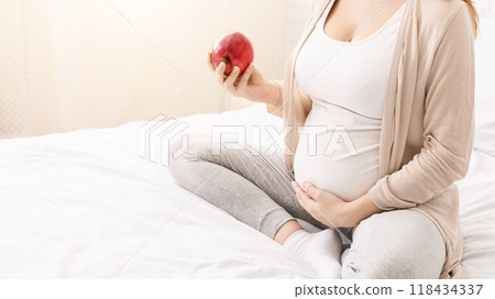 Cropped of pregnant woman is sitting on a bed, holding an apple in her hand. She appears to be relaxed and thoughtful, possibly reflecting on her pregnancy and health choices, copy space 118434337
