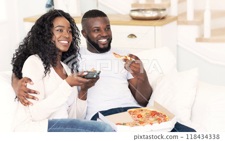 A couple relaxes on a white couch, watching TV and enjoying pizza. 118434338