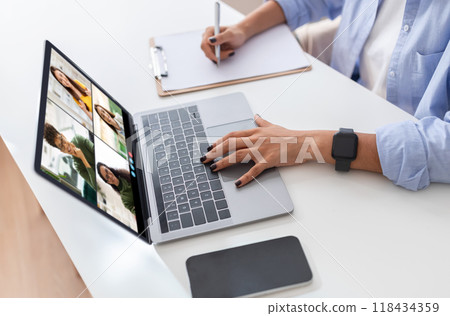 A woman is seated at a white desk with a laptop in front of her. She is using one hand to navigate the laptop while the other is resting on the desk. The laptop screen displays a video conference A woman is seated at a white desk with a laptop in front of her. She is using one hand to navigate the laptop while the other is resting on the desk. The laptop screen displays a video conference 118434359