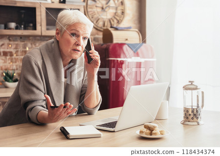 A senior woman with white hair sits at a kitchen table talking on a mobile phone while gesturing with her other hand. She is sitting in front of a laptop computer and a notebook. 118434374