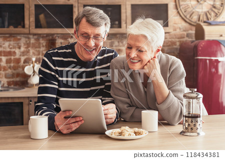A happy senior couple are sitting in their kitchen, looking at a tablet computer. The woman has a hand on her chin and is laughing, while the man is holding the tablet and smiling A happy senior couple are sitting in their kitchen, looking at a tablet computer. The woman has a hand on her chin and is laughing, while the man is holding the tablet and smiling 118434381