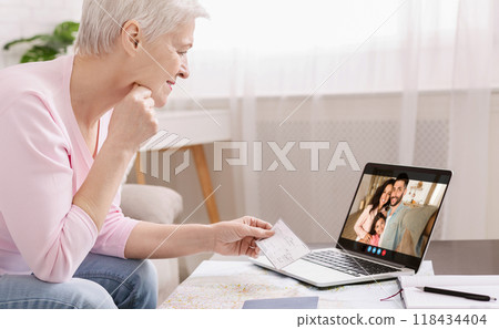 A senior woman is sitting at a table with a laptop in front of her. She is smiling and holding up a check for her family on the video call. The family is smiling back at her. A senior woman is sitting at a table with a laptop in front of her. She is smiling and holding up a check for her family on the video call. The family is smiling back at her. 118434404