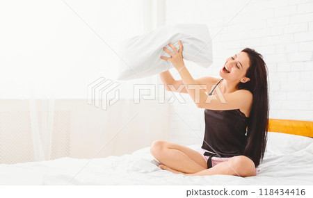 A woman is seen seated on a bed, clutching a pillow in her arms, singing. The room is minimally decorated, with only a bedside table visible in the background, copy space 118434416