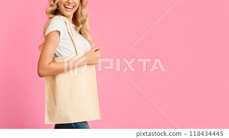 A woman with blonde hair and a bright smile is holding a blank canvas tote bag on a pink background. The image is well-lit and could be used for a variety of purposes, such as advertising, copy space A woman with blonde hair and a bright smile is holding a blank canvas tote bag on a pink background. The image is well-lit and could be used for a variety of purposes, such as advertising, copy space 118434445
