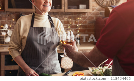 A senior woman in an apron stands in a kitchen and smiles as her partner toasts her with a glass of wine. The kitchen is decorated with exposed brick and a farmhouse style, cropped 118434465