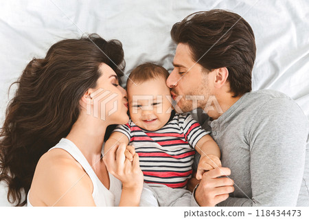 A happy family of three, a mother, a father, and a baby boy, are lying in bed together. The parents are each kissing the baby on either cheek as he smiles. The scene is full of love and affection. 118434473