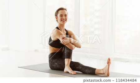 A cheerful young woman is seated on a yoga mat in a brightly lit room, engaging in a stretching exercise. She wears workout attire and has her hair tied back, showcasing a healthy, active lifestyle. A cheerful young woman is seated on a yoga mat in a brightly lit room, engaging in a stretching exercise. She wears workout attire and has her hair tied back, showcasing a healthy, active lifestyle. 118434484