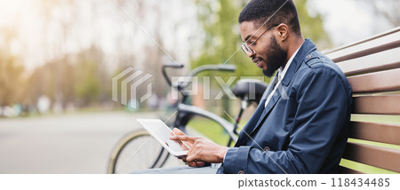 A professional black man is seated on a park bench, using a tablet with his bicycle next to him. The scene is peaceful and modern, highlighting a balance of technology and relaxation. 118434485