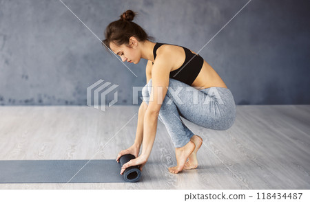A focused young woman is in the process of unrolling her yoga mat on a wooden floor in a tranquil studio setting, preparing for her exercise routine. 118434487