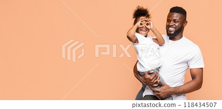 African American happy father and his little daughter smiling and playing together against a peach-colored backdrop. The father is holding his daughter in his arms, and she is playfully making glasses 118434507