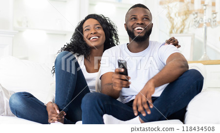 A happy couple sits on a couch, laughing and enjoying a movie or TV show together at home. 118434508