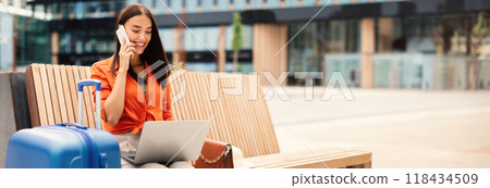Always In Touch. Happy Woman Traveler Talking On Mobile Phone While Working Online On Laptop Computer, Waiting For Flight Near Modern Airport Outside, Sitting With Suitcase, Copy Space 118434509