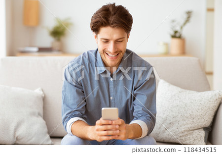 A man sits on a couch, smiling and looking at his phone. A man sits on a couch, smiling and looking at his phone. 118434515
