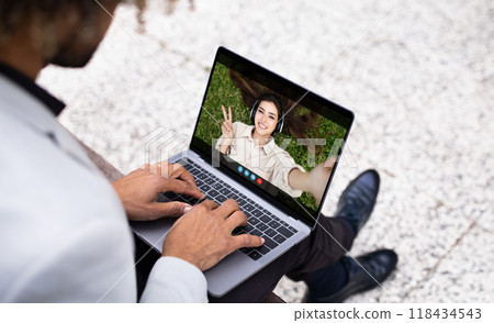 Black man sits outside on a bench, using a laptop to video chat with a woman. The woman is smiling and giving a peace sign. The man is typing on the laptop keyboard. 118434543