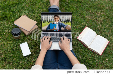 A student sits on the grass of a college campus, using a laptop to have a video call with another student. The student on the laptop screen is smiling and holding a book. 118434544