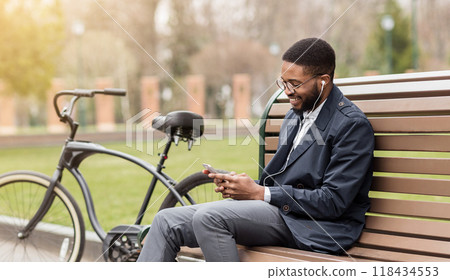 African American man is sitting on a park bench, engaged on his phone, with his bicycle nearby. The outdoor setting is calm and professional, indicating a blend of work and relaxation. African American man is sitting on a park bench, engaged on his phone, with his bicycle nearby. The outdoor setting is calm and professional, indicating a blend of work and relaxation. 118434553