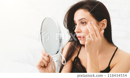 A woman is standing in front of a mirror, closely observing her reflection. She appears focused and intent as she examines her facial features and expressions closely. 118434554
