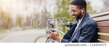 Black business professional is on his phone, sitting on a park bench with a bicycle next to him. The scene is calm and modern, blending work and leisure in a natural setting. 118434577