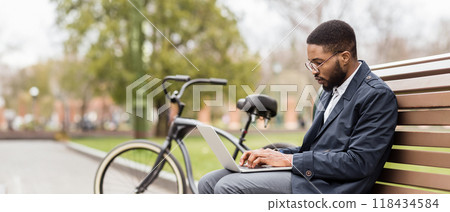 Black man with his laptop sits on a park bench, his bicycle close by. The setting is tranquil and professional, highlighting the balance between work and the outdoors. Black man with his laptop sits on a park bench, his bicycle close by. The setting is tranquil and professional, highlighting the balance between work and the outdoors. 118434584